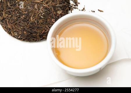Ciotola in porcellana Chawan con tè sheng pu-erh appena preparato. Torta di tè crudo in pu-erh pressata sul tavolo piena di foglie di tè invecchiate. Vero capolavoro artigianale Foto Stock