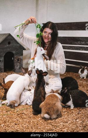 Donna in abito bianco siede in una penna di coniglio sulla segatura e nutre i conigli con la carota Foto Stock