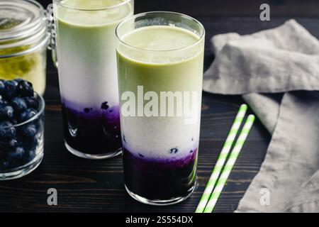 Lattine di Matcha al mirtillo ghiacciato in bicchieri alti: Lattine fredde a strati di tè verde, latte e mirtilli con cannucce di carta e ingredienti nelle vicinanze Foto Stock