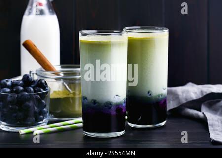 Lattine di Matcha al mirtillo ghiacciato in bicchieri alti: Lattine fredde a strati di tè verde, latte e mirtilli con cannucce di carta e ingredienti nelle vicinanze Foto Stock