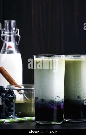 Lattine di Matcha al mirtillo ghiacciato in bicchieri alti: Lattine fredde a strati di tè verde, latte e mirtilli con cannucce di carta e ingredienti nelle vicinanze Foto Stock