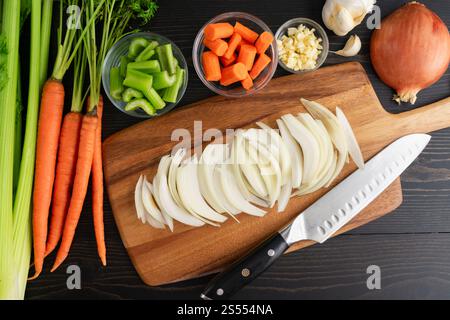 Cipolla gialla a fette su un tagliere di legno: Fette di cipolla su un tagliere con coltello santoku e verdure carote, sedano, aglio Foto Stock