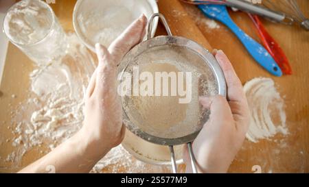 Vista dall'alto della giovane donna che scuote il setaccio per setacciare la farina mentre si prepara l'impasto. Vista dall'alto della giovane donna che scuote il setaccio per setacciare la farina mentre si prepara l'impasto Foto Stock