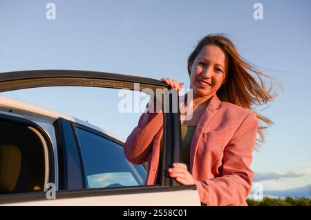 Donna d'affari sorridente che esce dalla sua auto durante un viaggio in auto al tramonto Foto Stock