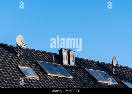 I pannelli solari sono installati sul tetto inclinato di una casa, completati da due parabole satellitari. Il cielo azzurro e limpido funge da sfondo, migliorando l'aspetto moderno della casa. Foto Stock