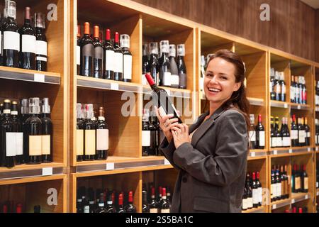 Allegra donna caucasica in giacca con una bottiglia di vino mentre fai shopping in un negozio di vini. Foto Stock