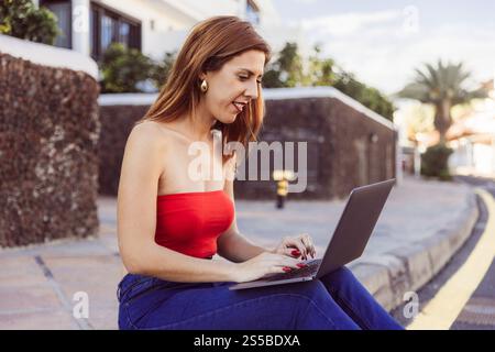 Una donna con la parte superiore rossa seduta su un marciapiede utilizza un notebook. Foto Stock