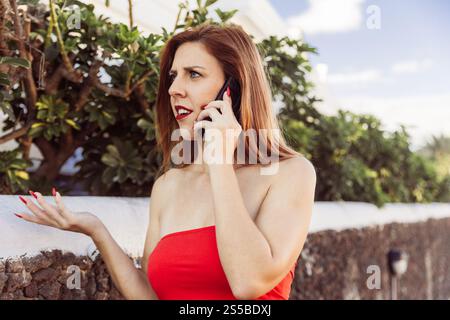 Una donna con un vestito rosso sta parlando al suo cellulare con un'espressione sorpresa. Foto Stock