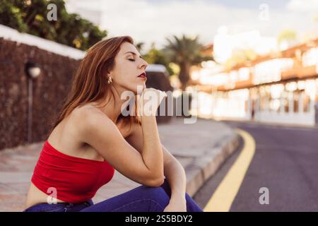 Una donna con il tetto rosso si siede sul marciapiede, occhi chiusi, look pensivo. Foto Stock