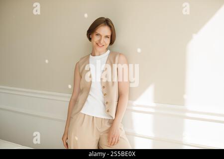 Giovane donna sorridente con un costume da pantaloni e un gilet appoggiati a una parete Foto Stock
