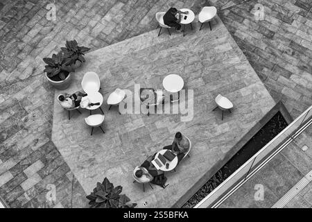 vista aerea di un'area salotto all'aperto con diversi tavoli rotondi e sedie bianche disposte su una superficie piastrellata. Poche persone sedute a usare un notebook Foto Stock