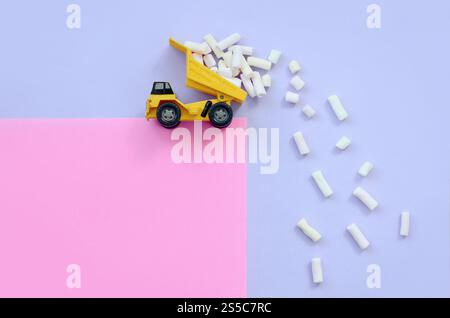 Il piccolo camioncino giallo lancia pezzi di marshmallow dalla schiena rialzata su uno sfondo color viola e rosa pastello. Vista dall'alto minima in posizione piatta. Scarico Foto Stock