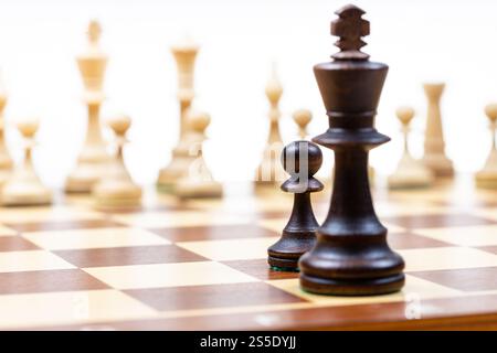 pedina nera e regina contro pezzi di scacchi bianchi sullo sfondo su scacchiera di legno primo piano (focus sulla pedina nera) Foto Stock