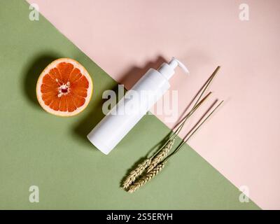 vista dall'alto di una bottiglia di plastica bianca con pompa vuota, metà di pompelmo fresco e orecchie essiccate naturali su sfondo di carta a colori Foto Stock