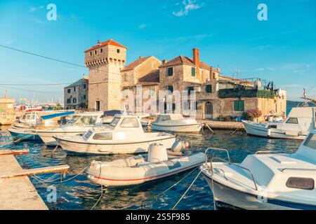 Piccole barche ormeggiate lungo un tranquillo canale con il monastero della fortezza di Kastel Gomilica e gli edifici tradizionali sotto un cielo azzurro, Kastela, Croazia Foto Stock