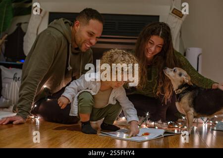 Una famiglia si diverte a stare insieme mentre un bambino dai capelli ricci legge un libro con luci a corda accese, accompagnato da un attento cane da compagnia, Ohio, USA Foto Stock