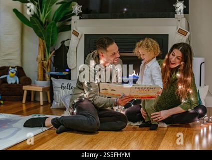 Una famiglia ama leggere un libro insieme accanto all'accogliente caminetto in un soggiorno illuminato da calore, Ohio, USA Foto Stock