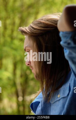 Profilo laterale di un uomo con capelli lunghi in una camicia blu, che guarda in lontananza all'aperto. Foto Stock