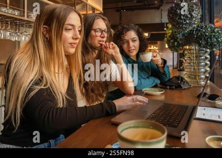 Tre donne si godono una conversazione e un drink in un accogliente tavolino, lavorano su un portatile, Berlino, Germania Foto Stock