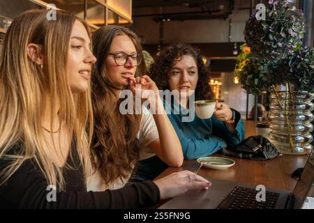 Tre donne si godono una conversazione e un drink in un accogliente tavolino, lavorano su un portatile, Berlino, Germania Foto Stock