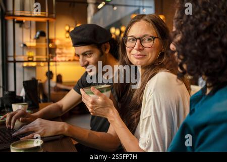 Tre amici o colleghi che condividono un momento in un accogliente bar, con un uomo che digita su un laptop e un'altra donna che si occupa di conversazione, Berlino, Germania Foto Stock