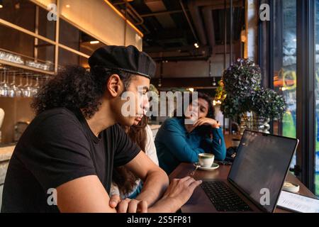 Un uomo con una camicia nera e un berretto lavora su un computer portatile in un bar con due donne, Berlino, Germania Foto Stock