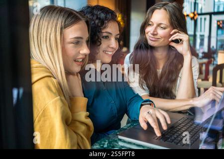 Tre donne sorridenti condividono un momento guardando insieme lo schermo di un laptop in un accogliente ambiente interno, Berlino, Germania Foto Stock