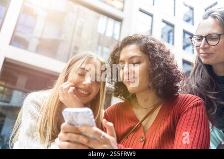 Tre donne condividono un momento di gioia guardando i contenuti su uno smartphone insieme all'aperto, Berlino, Germania Foto Stock