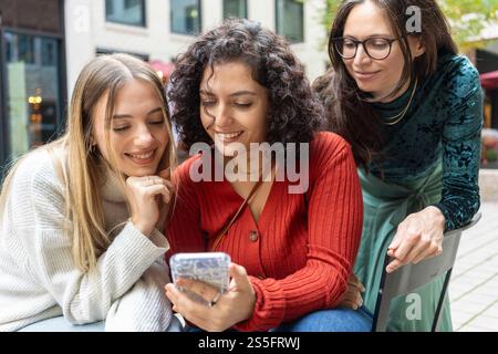 Tre donne condividono un momento guardando uno smartphone con curiosità e divertimento in una strada della città, Berlino, Germania Foto Stock