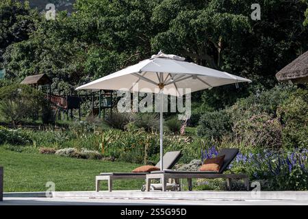 il parasole bianco proietta un'ombra su due poltrone imbottite. Sullo sfondo, un rigoglioso giardino verde si estende in lontananza, caratterizzato da vari p Foto Stock