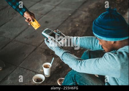 Mendicante seduto sotto il cavalcavia con una macchina per carte di credito e carte di credito. Foto Stock