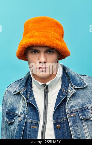 Il bellissimo giovane presenta un elegante cappello arancione, vestito in denim, su uno sfondo blu brillante. Foto Stock