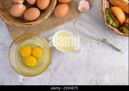 Vista dall'alto con tuorlo nel recipiente di vetro e nel cestino di vimini, hamburger e hotdog con fette di pomodoro e lattuga nel cestello per la tessitura, olio in tazza e frusta per uova Foto Stock