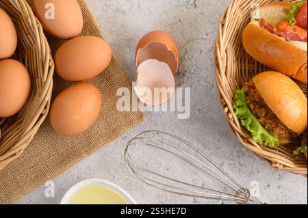 Vista dall'alto con tuorlo nel recipiente di vetro e nel cestino di vimini, hamburger e hotdog con fette di pomodoro e lattuga nel cestello per la tessitura, olio in tazza e frusta per uova Foto Stock