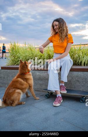 La giovane donna che indossa un abbigliamento casual è seduta su un confine di cemento e sta addestrando il suo cane careliano con dolcetti, il suo longboard poggia a terra accanto ai suoi piedi Foto Stock