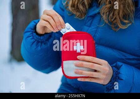 Primo piano delle mani di una donna che prende pillole da un kit di pronto soccorso rosso con sopra una croce bianca. In piedi in una foresta invernale innevata. Sottolineare l'importanza della preparazione per le attività all'aperto Foto Stock