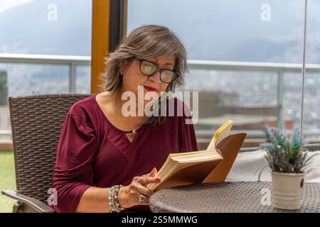 Ritratto di donna anziana con occhiali e camicetta Bordeaux che legge un libro a un tavolo di rattan all'aperto Foto Stock