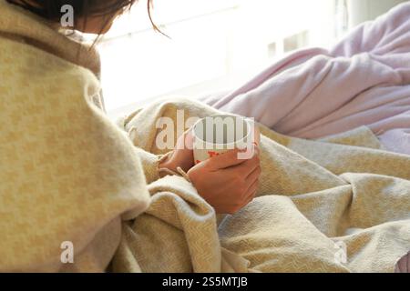 Una giovane donna con un raffreddore a letto a casa, avvolta in una coperta e tenendo una tazza di bevanda calda con entrambe le mani Foto Stock