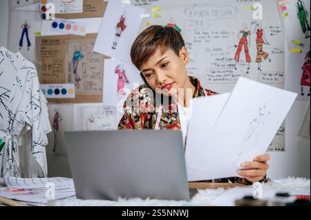 Un giovane stilista tiene in mano la carta da schizzo di moda e parla con lo smartphone mentre utilizza il computer portatile per cercare idee per abiti di design e nastri Foto Stock