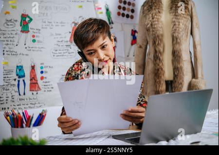 Un giovane stilista tiene in mano la carta da schizzo di moda e parla con lo smartphone mentre utilizza il computer portatile per cercare idee per abiti di design e nastri Foto Stock