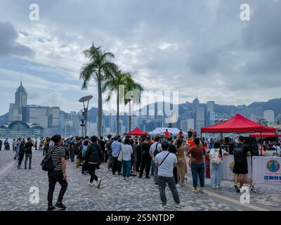 Hong Kong - 16 novembre 2024 : Un vivace mercato all'aperto si svolge sotto un cielo nuvoloso, con palme che incorniciano uno sfondo urbano. Molte persone navigano Foto Stock