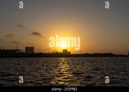 Uno splendido tramonto dorato che diffonde calde sfumature sullo skyline della città costiera. Il riflesso scintillante sull'acqua increspata aggiunge profondità e tranquillità Foto Stock