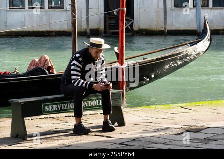 Un gondoliere si siede con il suo telefono vicino a un canale a Venezia, in Italia. Foto Stock