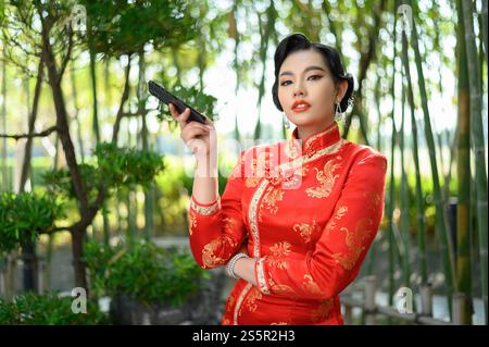 Ritratto bella donna asiatica in un cheongsam cinese in posa con smartphone su sfondo foresta di bambù, spazio copia, concetto di Capodanno cinese Foto Stock