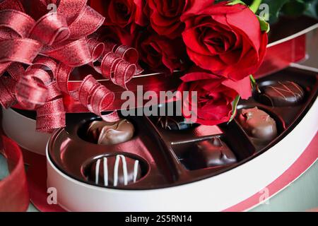Bellissime rose rosse a gambo lungo con una scatola a forma di cuore di caramelle al cioccolato miste. Messa a fuoco selettiva sui pezzi centrali delle caramelle. Sfondo sfocato. Foto Stock