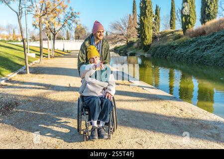 Giovane che assiste una donna anziana in sedia a rotelle che indica qualcosa mentre cammina in un parco vicino a un fiume o a un lago Foto Stock