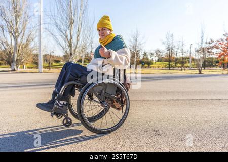 Uomo felice su sedia a rotelle che mostra i pollici mentre si gode una giornata di sole nel parco, promuovendo attività ricreative accessibili e un atteggiamento positivo verso la disabilita' Foto Stock