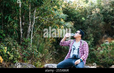 giovane uomo attraente con zaino da viaggio e macchina fotografica seduti per riposare sulle rocce nella foresta di montagna e bere l'acqua dalla bottiglia e copiare lo spazio Foto Stock