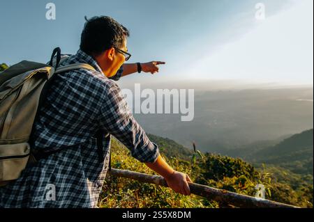 Giovane escursionista asiatico con zaino in piedi e punta il dito in avanti verso la splendida vista della valle al punto panoramico con raggi del sole al tramonto, copia spazio Foto Stock
