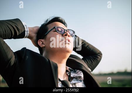 Il giovane uomo d'affari asiatico che indossa un abito con cravatta e occhiali chiude gli occhi stanchi e dorme per rilassarsi all'aperto Foto Stock